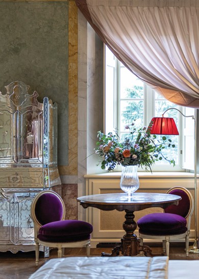 A luxury Italian hotel room features purple velvet chairs and a table set with a floral arrangement. A glass wardrobe stands between curtained windows.