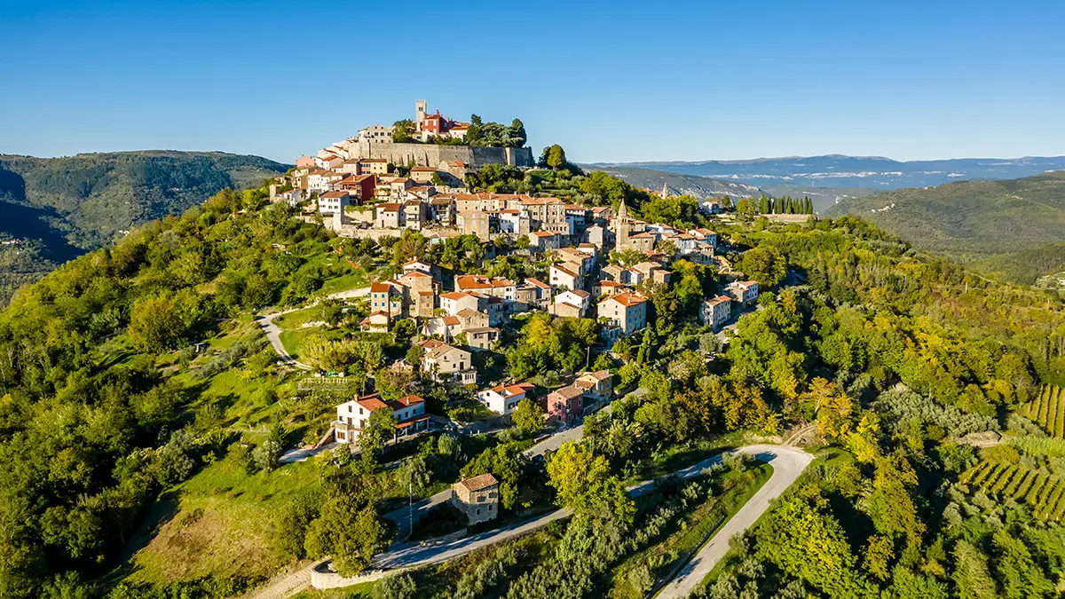 Medieval buildings sit on a lush green hillside surrounded by a winding road in Croatia's Istria region.